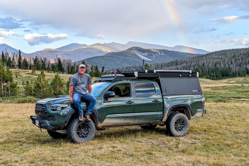 Dave Lalande's Toyota Tacoma with a pop-up camper and a Starlink dish on the roof, parked in a mountain meadow under a rainbow.